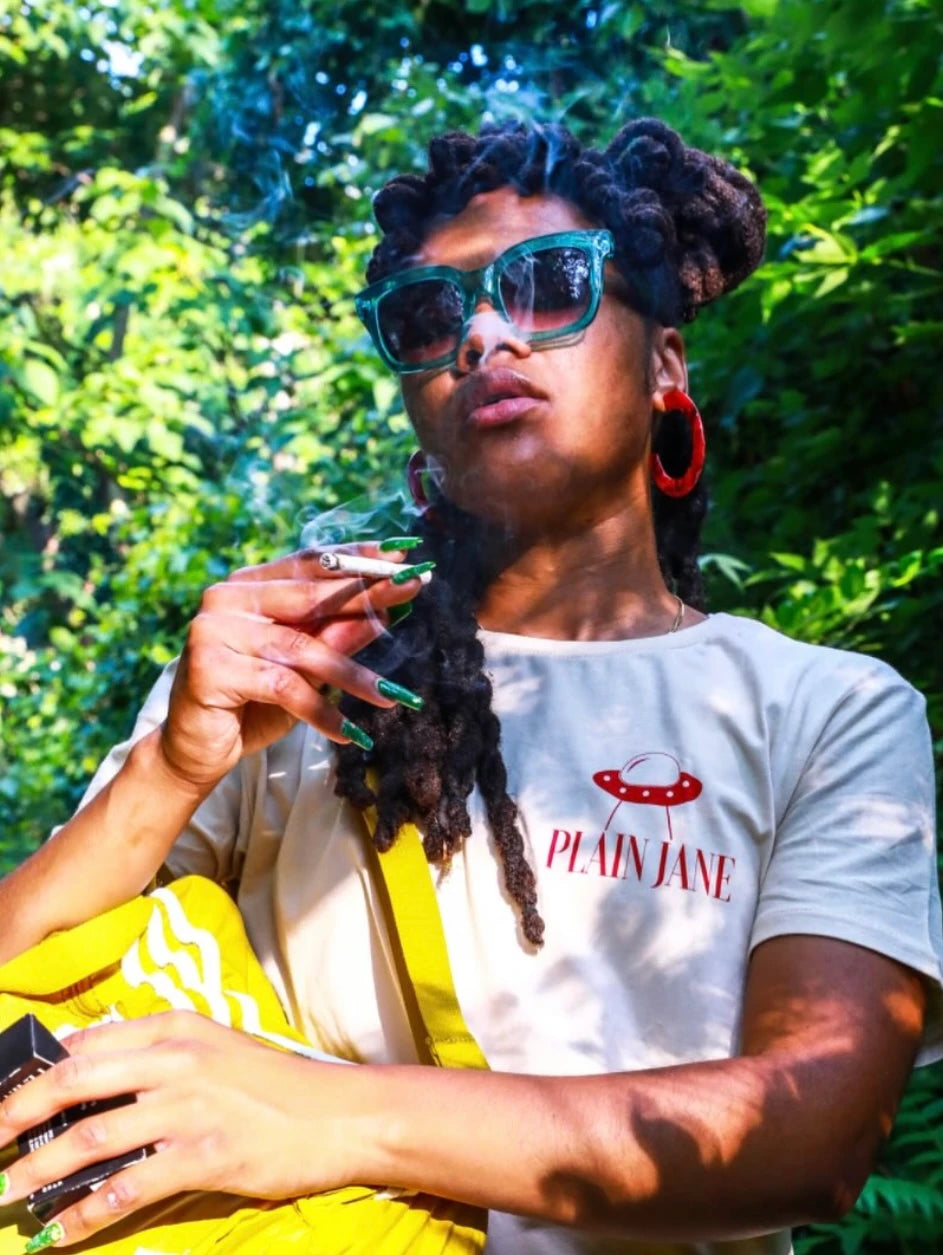 A woman outdoors wearing teal sunglasses and a white shirt with the red Plain Jane wordmark and UFO graphic