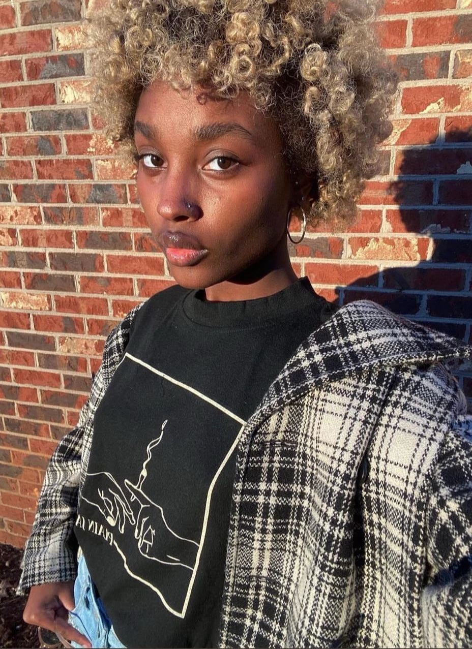 A woman wearing a black Plain Jane shirt and plaid jacket against a brick wall